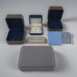 May include: An assortment of jewelry boxes in various shapes and sizes. The boxes are in shades of gray, blue, and brown, with soft velvet interiors. Some boxes are open, revealing their interior. A small rectangular box displays the text "Mia Monserrat".