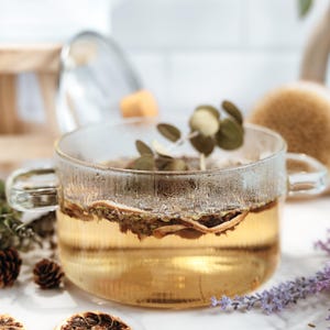 Könnte beinhalten: Eine transparente Glastasse gefüllt mit goldenem Tee, Kräutern und getrockneten Zitrusfrüchten. Die Tasse hat einen Henkel und steht auf einer weißen Oberfläche, umgeben von getrockneten Zitronenscheiben, Tannenzapfen und Lavendelzweigen. Eine natürliche, spa-ähnliche Ästhetik.
