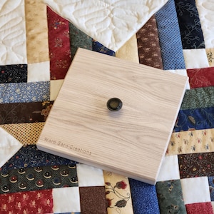 May include: A light-colored wooden square with a dark knob in the center, resting on a patchwork quilt. The quilt features various fabric patterns in red, blue, and cream. The text "Ward Barn Creations" is visible on the wood.