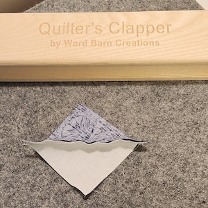 Peut inclure: Un outil de quilting en bois appel&eacute; "Quilter's Clapper" de Ward Barn Creations. L'outil est utilis&eacute; pour presser le tissu &agrave; plat pendant le quilting. Un morceau de tissu avec un motif bleu et blanc est montr&eacute; au premier plan.