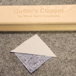 Peut inclure: Un outil de quilting en bois appel&eacute; "Quilter's Clapper" de Ward Barn Creations. L'outil sert &agrave; presser le tissu &agrave; plat. Un carr&eacute; de tissu blanc et bleu est pr&eacute;sent&eacute; sous l'outil.