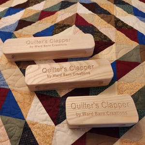 Peut inclure: Trois clapets de matelassage en bois avec le texte "Quilter's Clapper by Ward Barn Creations" grav&eacute; sur chacun. Les clapets sont sur une courtepointe patchwork color&eacute;e.