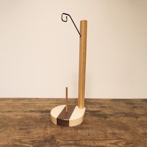 May include: A wooden paper towel holder with a dark metal hook. The base is a light wood with a dark wood accent. The holder has a tall, vertical wooden post and a smaller wooden dowel.