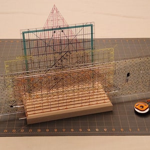 Puede incluir: Un soporte de madera con cuatro reglas de pl&aacute;stico transparente apiladas encima. Las reglas est&aacute;n marcadas en pulgadas y tienen un patr&oacute;n de cuadr&iacute;cula. Un cortador rotatorio con un mango naranja est&aacute; sobre una estera de corte gris.