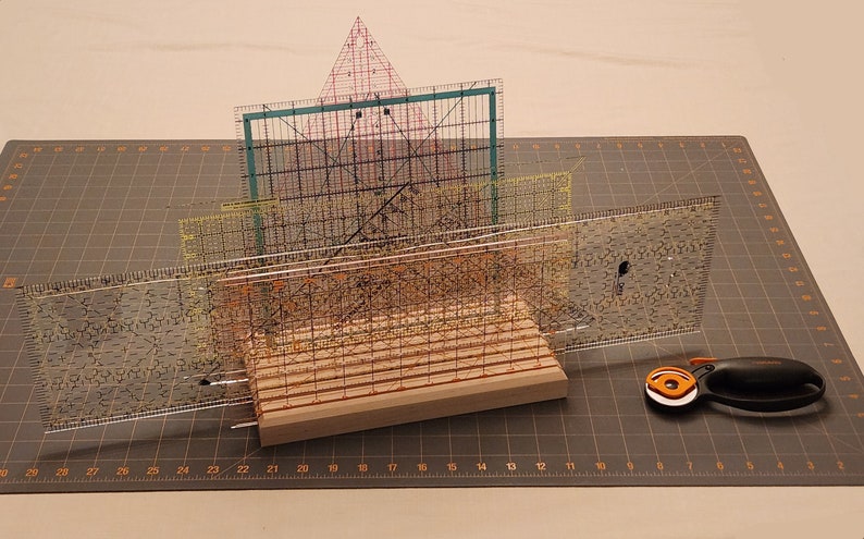 Puede incluir: Un soporte de madera con cuatro reglas de pl&aacute;stico transparente apiladas encima. Las reglas est&aacute;n marcadas en pulgadas y tienen un patr&oacute;n de cuadr&iacute;cula. Un cortador rotatorio con un mango naranja est&aacute; sobre una estera de corte gris.