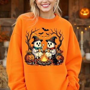 May include: Orange sweatshirt featuring a Halloween-themed graphic. The design shows two cartoon ghosts wearing witch hats, sitting by a campfire, with pumpkins, a black cat, and bats. The ghosts are holding mugs. The background includes trees and string lights.