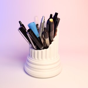 May include: A white ceramic pen holder shaped like a column, filled with a variety of pens and markers, including black, blue, and yellow.