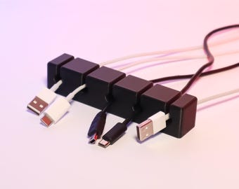 May include: Black cable organizer with six slots to hold and organize cords.  The organizer is made of plastic and has a matte finish.  The organizer is holding a variety of cords, including USB cables, charging cables, and audio cables.
