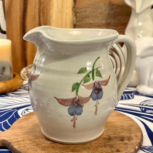 May include: A white ceramic pitcher with a floral design. The pitcher has a wide mouth and a handle. The flowers are painted in shades of blue, purple, and green.