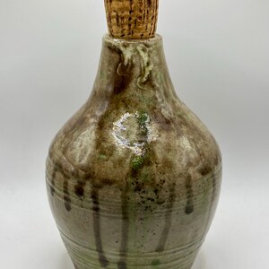 May include: A handcrafted ceramic bottle with a cork stopper. The bottle features a unique design with shades of brown, green, and tan, with a dripped glaze effect. The bottle has a rounded shape and a textured surface.