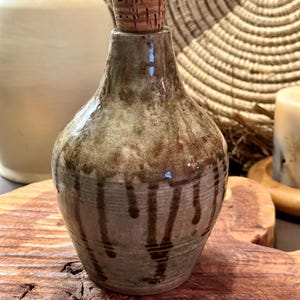 May include: A small, hand-thrown ceramic bottle with a cork stopper. The bottle has a light brown glaze with dark brown streaks and drips. It is sitting on a wooden surface.