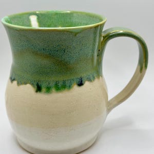 May include: A handmade ceramic mug with a green interior and rim, transitioning to a light green and beige exterior. The mug has a rounded body, a sturdy handle, and a white base.