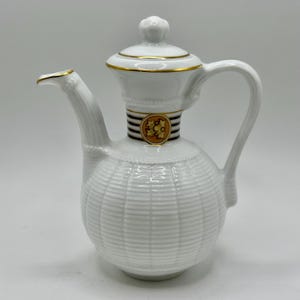 May include: White ceramic coffee pot with a lid, handle, and spout. The pot features gold trim and a decorative band with a gold emblem. The body has a textured, woven pattern.