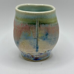 May include: A handmade ceramic mug with a unique, colorful glaze. The mug features a rounded body and a slightly flared rim. The glaze includes shades of blue, pink, green, and yellow, creating a textured, artistic appearance. A vertical crack runs down the side.