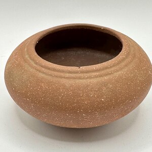 May include: A small, round, terracotta-colored ceramic bowl with a wide opening. The bowl has a textured surface with small white speckles and a slightly raised rim. The interior is a darker shade of brown.