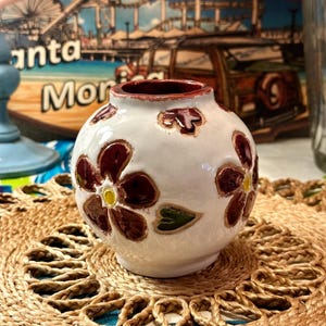 Puede incluir: Maceta de cerámica blanca con borde marrón y flores rojas pintadas con centros amarillos. La maceta está sobre un mantel tejido marrón.