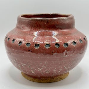 May include: A handmade, reddish-brown ceramic vase with a rounded body and a short, cylindrical neck. The vase features a series of small, dark blue holes around the upper portion and sits on a light brown base. The surface has a slightly textured appearance.