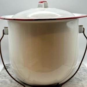 May include: A white enamel bucket with a matching lid, featuring a thin red rim and a red accent on the lid handle. The bucket includes a dark brown metal wire handle with a black grip, and the overall design is a cylindrical container.