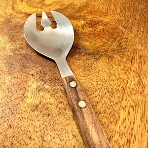 May include: A stainless steel salad server with a wooden handle. The server has a wide, slotted head with a central point and two side tines. The wooden handle is attached with two brass rivets, and the server rests on a wooden cutting board.
