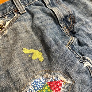 May include: Blue denim jeans with a patchwork design on the bottom right corner. The patchwork is made of colorful fabric with floral and geometric patterns. There is a yellow paint splatter on the left side of the jeans.