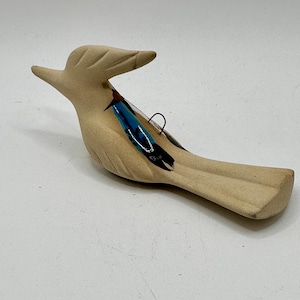 May include: A tan-colored bird figurine with a long body and a curved beak. The bird has a blue and black feather detail on its side. The figurine is made of a light-colored material and is likely a decorative item.