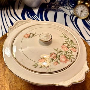 May include: A white ceramic serving dish with a lid, featuring a floral design of pink roses and green leaves. The dish has a gold trim around the edge and a gold knob on the lid.