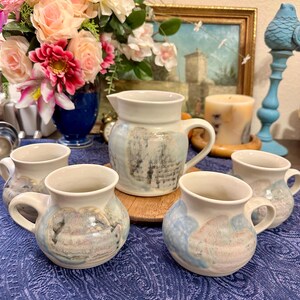 May include: A set of five ceramic mugs and a pitcher with a blue and white glaze. The mugs have a rounded shape and a handle. The pitcher has a wider base and a spout. The mugs and pitcher have a rustic, hand-painted look.