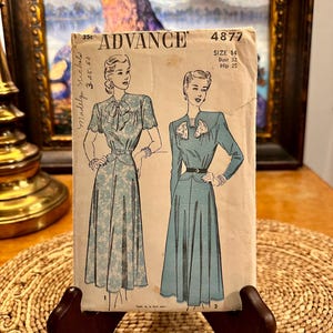 May include: Vintage sewing pattern for a dress, Advance pattern number 4877, size 14, bust 32, hip 35. The pattern shows a woman wearing a blue dress with a white floral print and a white bow at the neck.