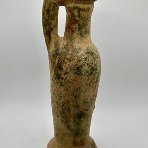 May include: A weathered, antique-style amphora with a single handle. The ceramic vessel features a rounded base and a narrow neck. The surface is textured with shades of brown, green, and tan, suggesting age and use. A decorative piece.