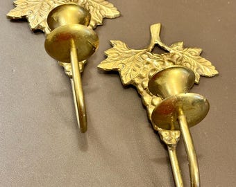May include: Two vintage brass wall sconces. Each sconce features a grape leaf design with a candle holder. The sconces are made of brass and have a warm, golden color. The design is detailed and adds a touch of elegance.