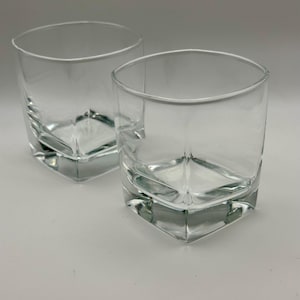 May include: Two clear, square-shaped drinking glasses. The glasses have a thick, geometric base and a slightly curved rim. The glasses are empty and appear to be made of glass. The background is a solid, light gray color.