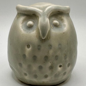 May include: A ceramic owl figurine with a light gray glaze and a speckled texture. The owl has large, round eyes and a prominent beak.