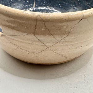 May include: A white ceramic bowl with a blue glaze inside. The bowl has a crackled finish and a small hole on the side.