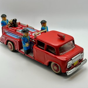 May include: A vintage red toy fire truck with blue and white accents. The toy features three firefighter figures in blue uniforms and black hats. The truck has white-rimmed wheels and a silver front bumper.