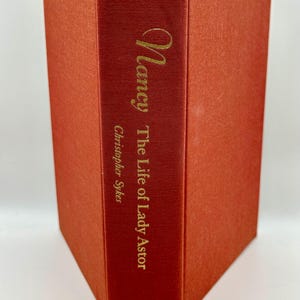 May include: A hardcover book titled "Nancy The Life of Lady Astor" by Christopher Sykes. The book has a textured, reddish-orange cover and spine. The title and author's name are in gold lettering. The publisher's name, Harper & Row, is at the bottom.