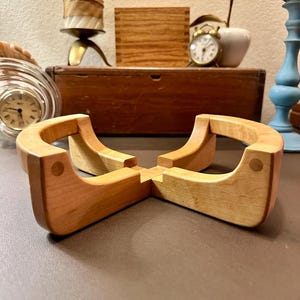 May include: A light-colored wooden trivet with a four-part design. Each part curves upward, creating a circular shape when assembled. The trivet is made of light-colored wood and has a smooth finish. Other items in the background include a candle holder, a small clock, and a wooden box.