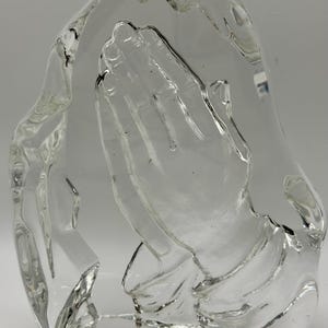 May include: A clear glass sculpture depicting a pair of hands clasped in prayer. The hands are detailed, with visible fingers and a textured surface. The sculpture is set against a plain white background.