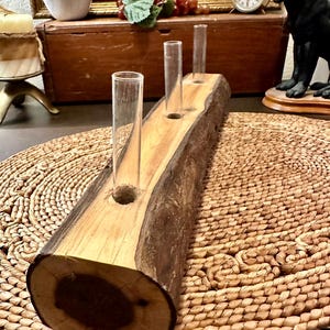 May include: A wooden centerpiece with three glass test tubes. The wood has a natural edge and a light brown color with darker brown bark. The test tubes are clear and cylindrical, perfect for displaying small flowers or stems.