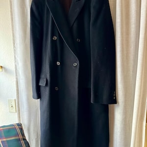 May include: A long, black double-breasted coat with a notched lapel and six buttons. The coat is hanging on a wooden hanger against a neutral background. The coat has a classic, tailored design.