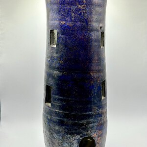 Puede incluir: Una torre de cerámica azul con detalles blancos y ventanas recortadas. La torre tiene una superficie texturizada y una base redondeada.