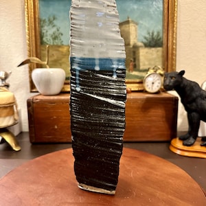 May include: A tall, rectangular ceramic vase with a textured surface. The top section is light gray, transitioning to a blue band, and then to a black base. White drips cascade down the black portion. The vase is approximately 12 inches tall.
