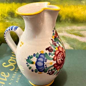 May include: White ceramic pitcher with a yellow rim and a floral design in shades of blue, red, green, and yellow. The pitcher has a handle and a spout.