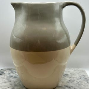 May include: A ceramic pitcher featuring a two-tone design with a glossy grey upper section and a smooth cream-colored lower section. The vessel has a curved handle on the right side and a small pouring spout at the top rim, with a rounded body.