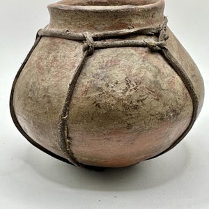 May include: A rustic, hand-crafted pottery vessel with a rounded body and a slightly flared rim. The pot is a blend of earthy tones, with a reddish-brown base and a mottled, textured surface. A leather-like material is wrapped around the pot.