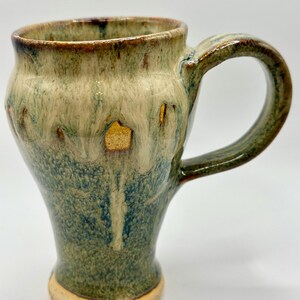 May include: Handmade ceramic mug with a unique, organic shape. The mug features a blend of green, blue, and tan glazes, creating a marbled effect. The handle is rounded and the base is a lighter tan color. A functional and decorative piece.