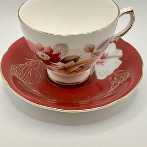 May include: A vintage teacup and saucer set with a floral design. The cup and saucer are white with a gold trim. The floral design features pink and red roses with green leaves. The saucer is a deep red color.