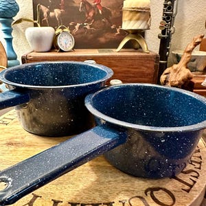 May include: Two blue speckled enamelware pots with handles sit on a wooden cutting board. The pots are dark blue with white speckles. The cutting board has the word "QUALITY" engraved on it. A vintage painting and other decorative items are in the background.