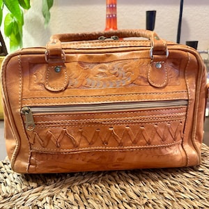 May include: A tan leather handbag with a top handle and a zipper closure. The bag features embossed floral and geometric patterns. The bag is sitting on a woven surface.