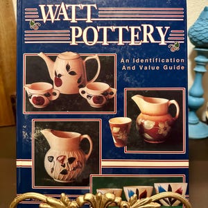 May include: A book titled "WATT POTTERY" with images of pottery pieces. The cover is dark blue with red and white text and features a teapot, creamers, and a pitcher. The book is an identification and value guide.