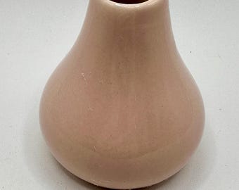 May include: A small, pear-shaped vase in a soft, muted pink. The ceramic vase has a rounded base that tapers to a narrow neck with a circular opening. The smooth surface reflects light, highlighting its simple, elegant form. A decorative item.